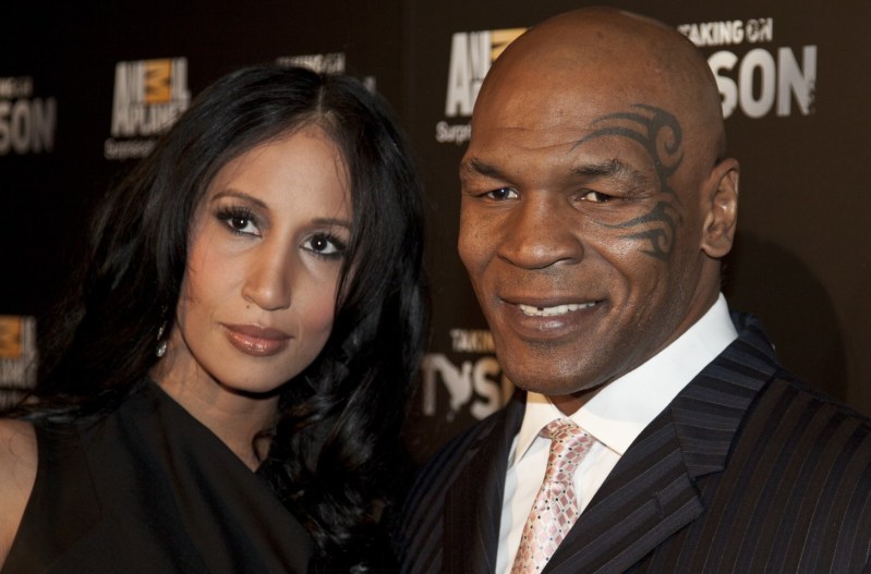 Mike Tyson and his wife, Lakiha Spicer, are standing close and smiling at the camera. Mike is wearing a black, striped suit, and Lakiha has dark curly hair and is wearing a black dress.