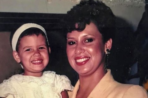 Victoria Eugenia Henao is smiling and holding a baby, Manuela Escobar, who is grinning and wearing a white dress and headband.
