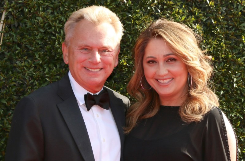 Pat Sajak and his wife, Lesly Brown, wearing black tie attire and standing in front of a green backdrop.