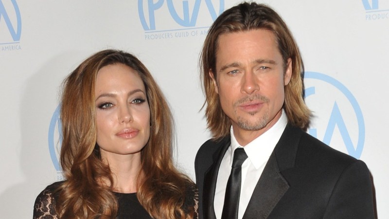 Angelina Jolie, in a black dress, stands with Brad Pitt, in a black suit, in front of a white background