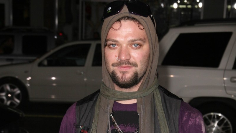 Bam Margera wears a purple t shirt under a brown hoodie on the street at night