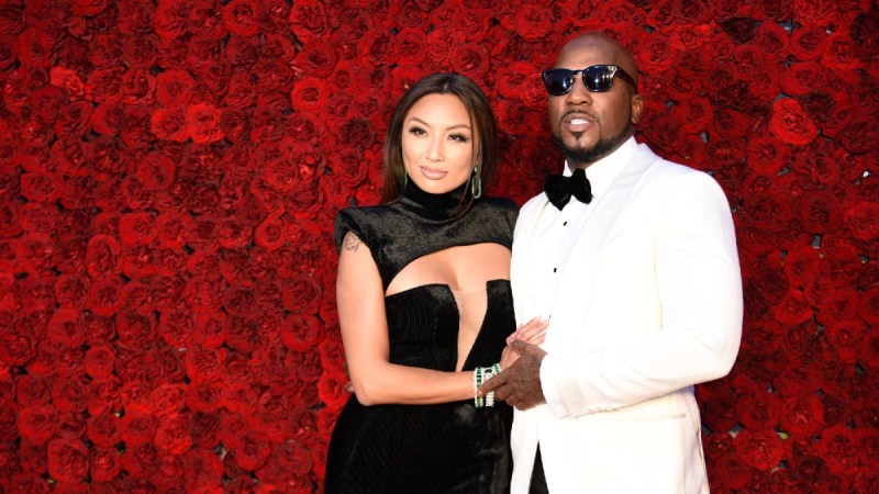 Jeannie Mai, in a black dress, stands with Jeezy, in a white tux, against a red floral background