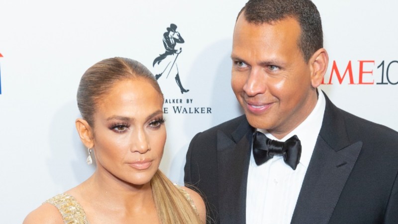 Jennifer Lopez, in a gold dress, stands with Alex Rodriguez, in a black tux, before a white background