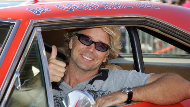 John Schneider wears a gray shirt and sits in the iconic Dukes Of Hazzard car