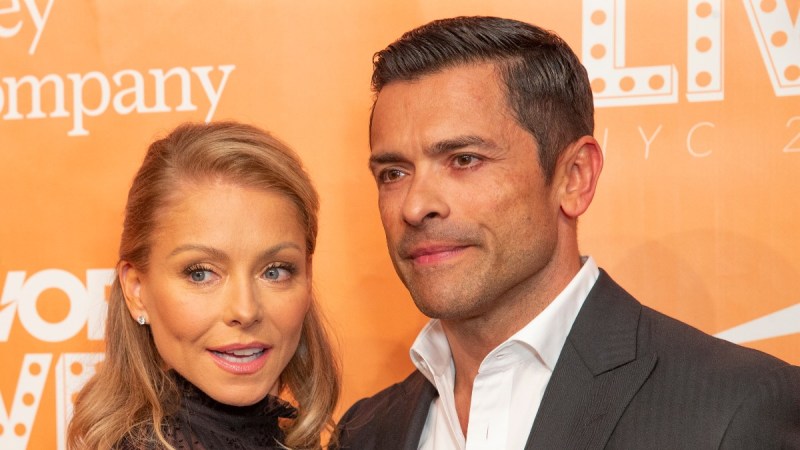 Kelly Ripa, in a black dress, cozies up to Mark Consuelos, in a dark gray suit, against an orange background