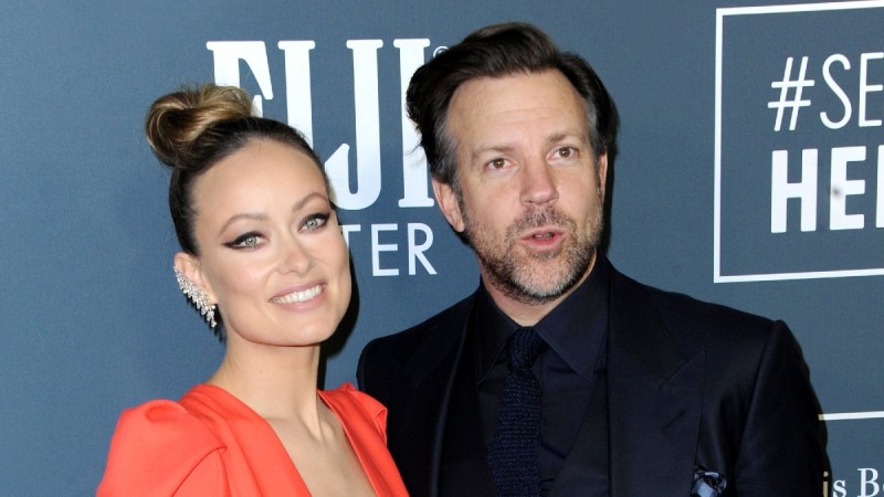 Olivia Wilde, in an orange dress, stands with Jason Sudeikis, in a black suit, on the red carpet