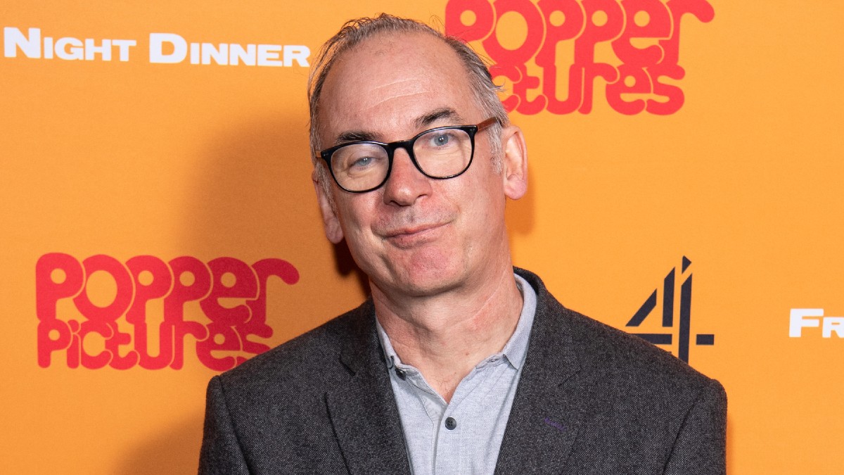 Paul Ritter wears a gray suit jacket against a bright orange background