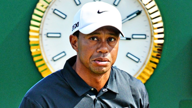 Tiger Woods wears a black golf shirt and white hat in front of a clock