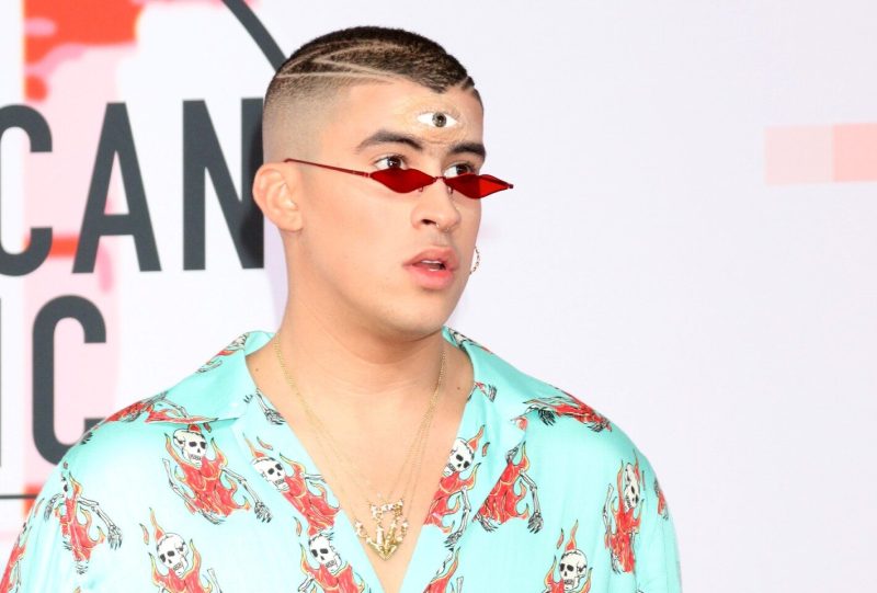 Bad Bunny wearing red sunglasses and a blue shirt with skulls printed on it. There's a prosthetic third eye on his forehead.