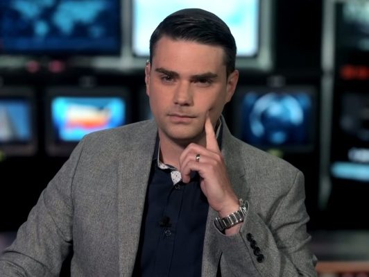 Ben Shapiro resting his finger on his face and wearing a grey suit.