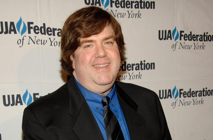 Dan Schneider smiling at a red carpet event and wearing a blue button-down shirt with black jacket