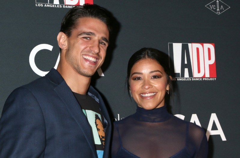 Gina Rodriguez and her husband, Joe LoCicero, smiling at a red carpet event. He is wearing a blue jacket and she is wearing a sheer navy blue dress.