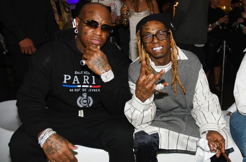 Birdman wearing a black track suit sitting with Lil Wayne who is smiling and wearing a white striped jacket.