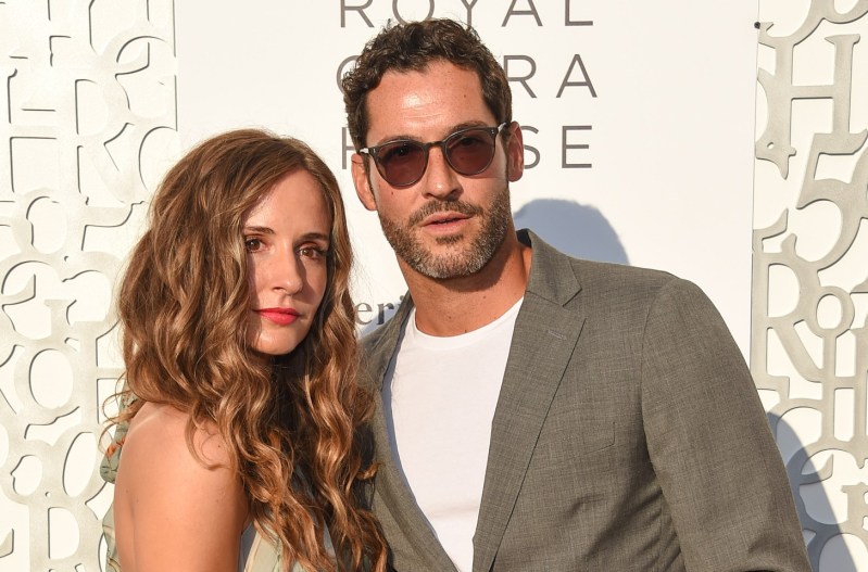 Tom Ellis in a grey suit with his wife, Meaghan Oppenheimer, who is wearing a green and beige dress.