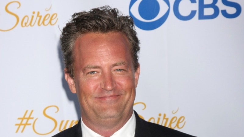 Matthew Perry wears a black suit against a white background on the red carpet