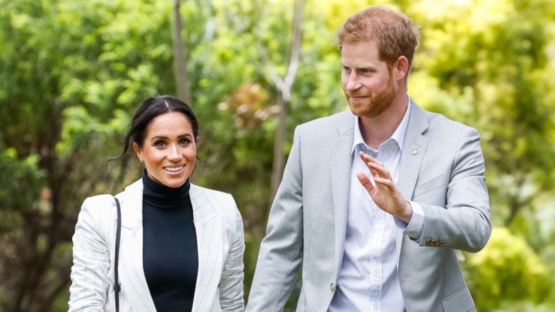 Meghan Markle, in a dark top and white blazer, walks hand in hand with Prince Harry, in a white shirt and gray blazer