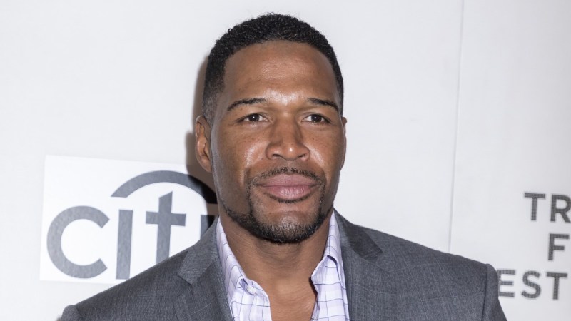 Michael Strahan wears a gray suit against a white background with grey lettering
