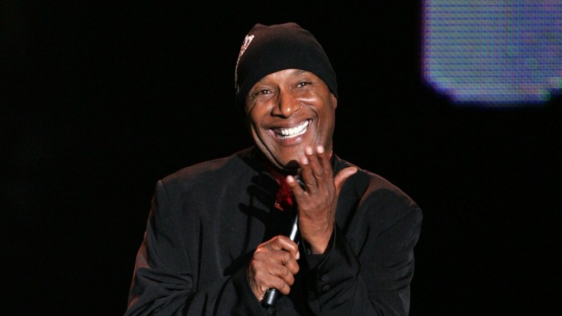 Paul Mooney smiles on stage while wearing a black jacket