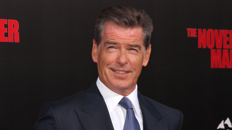 Pierce Brosnan wears a dark suit while posing in front of a black background with red lettering