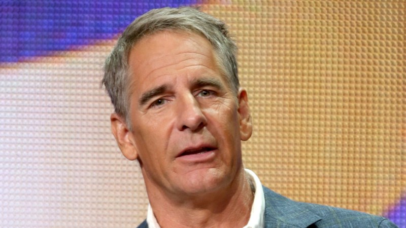 Scott Bakula wears a suit onstage against an orange and purple background