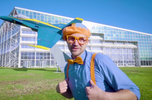 Blippi wearing a blue short, orange suspenders, orange bow tie, and a matching beret while standing in front of a blue airplane.