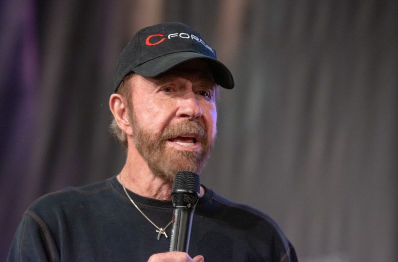 Chuck Norris wearing a black sweater and black hat, speaking into a microphone.