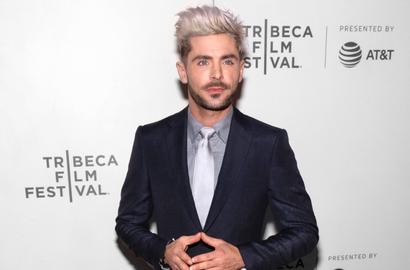 Zac Efron holding his hands out and wearing a navy blue suit.