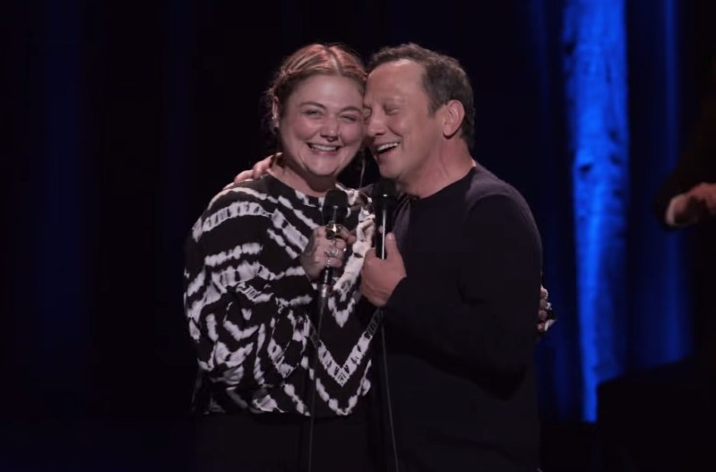 Elle King in a black and white shirt hugging Rob Schneider on stage, who is wearing a black shirt.