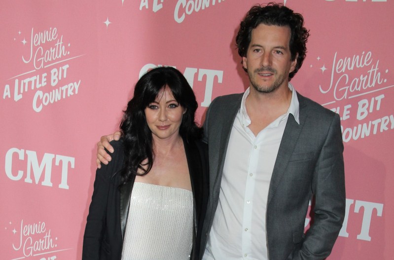 Shannen Doherty and spouse Kurt Iswarienko at a CMT event. She's wearing a black jacket and a white shirt; he has his arm around Doherty and is wearing a grey jacket and white shirt.