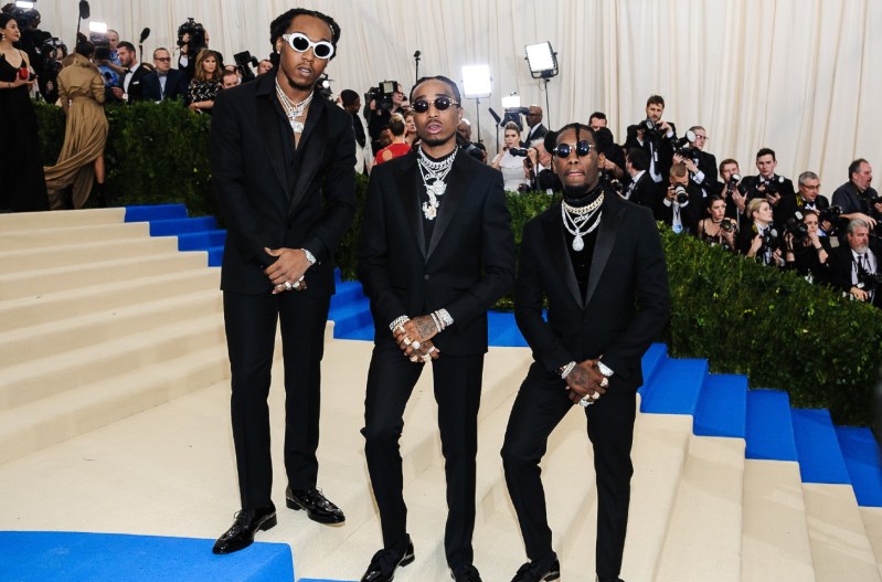 Migos at the Met Gala wearing matching black suits standing on steps in front of a bunch of photographers.