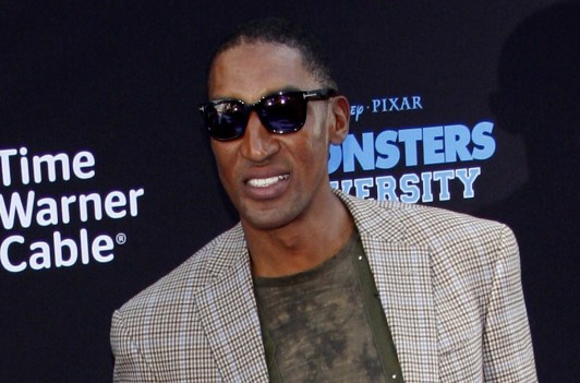 Scottie Pippen smiling and wearing sunglasses with a green shirt and brown plaid jacket.