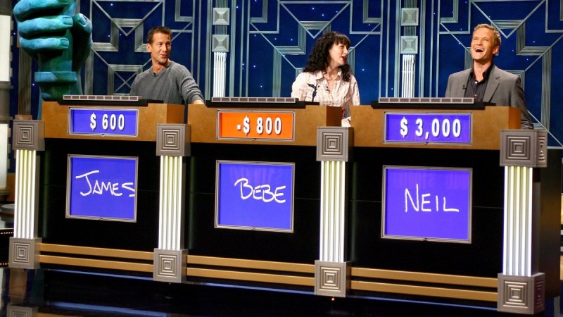 Three celebrity contestants on Jeopardy! laugh together behind their individual podiums
