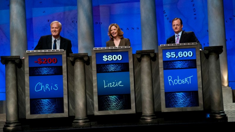 Three people stand behind podiums on the set of Jeopardy!