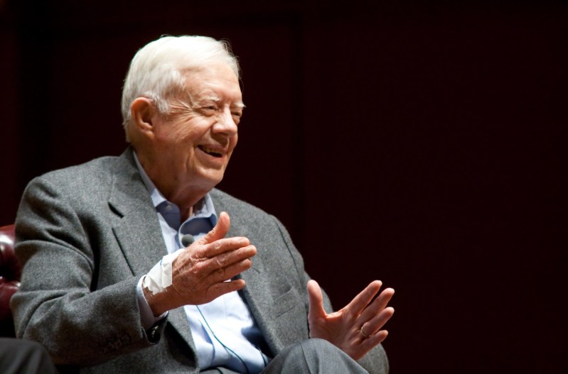 Jimmy Carter sitting in a grey suit and holding his hands out while giving a speech.