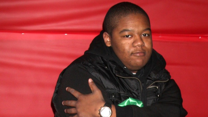 Kyle Massey wears a black jacket and crosses his arms in front of a red backdrop