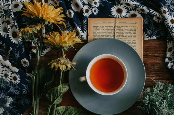 Image of a cup of tea on top of a book and next to some flowers.