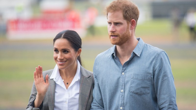 Meghan Markle wears a gray jacket and waves as Prince Harry walks close by in a blue shirt