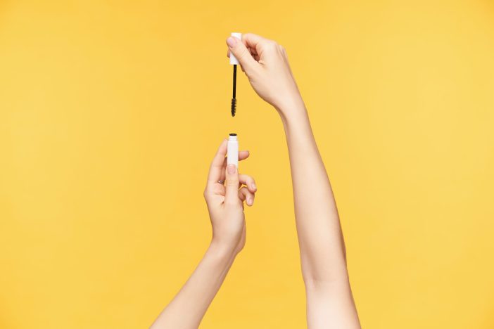 Image of woman opening mascara against a yellow background.
