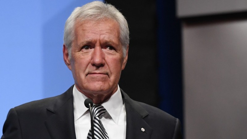 Alex Trebek wears a dark suit against a blue and black background