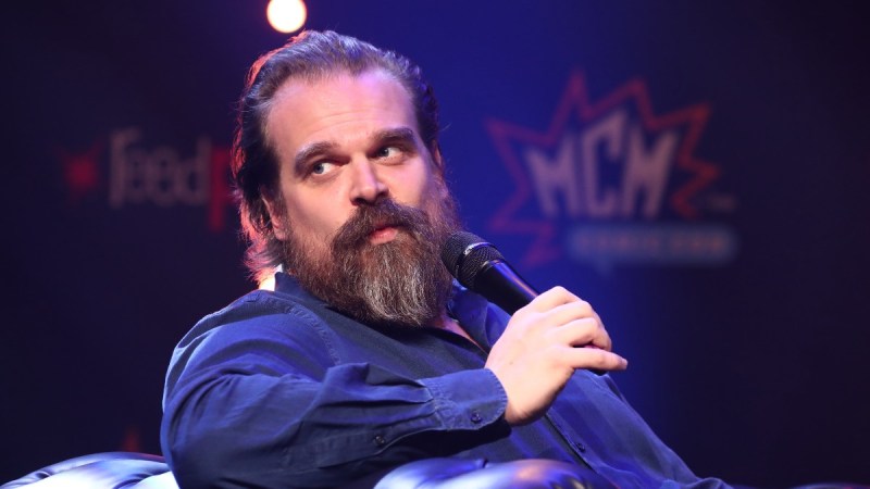 David Harbour wears a dark shirt and leans back as he holds a microphone onstage