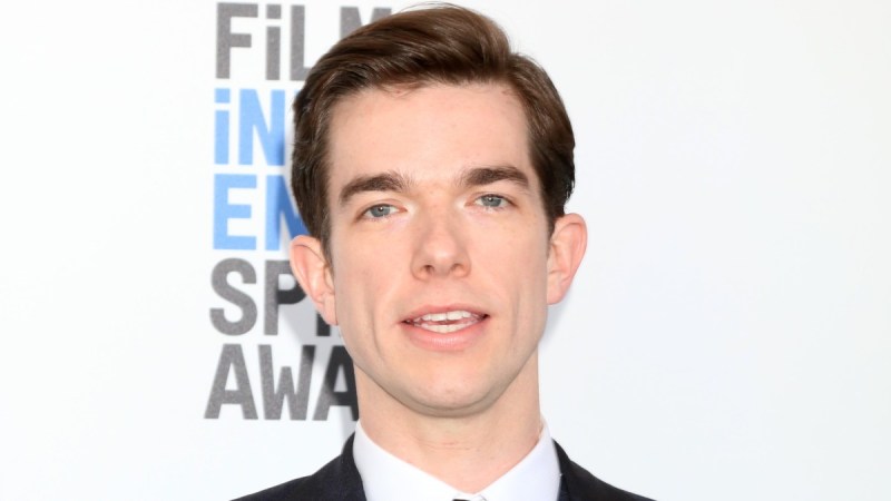 John Mulaney wears a dark suit against a white background on the red carpet