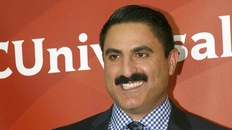 Reza Farahan wears a black suit against a deeep orange background on the red carpet