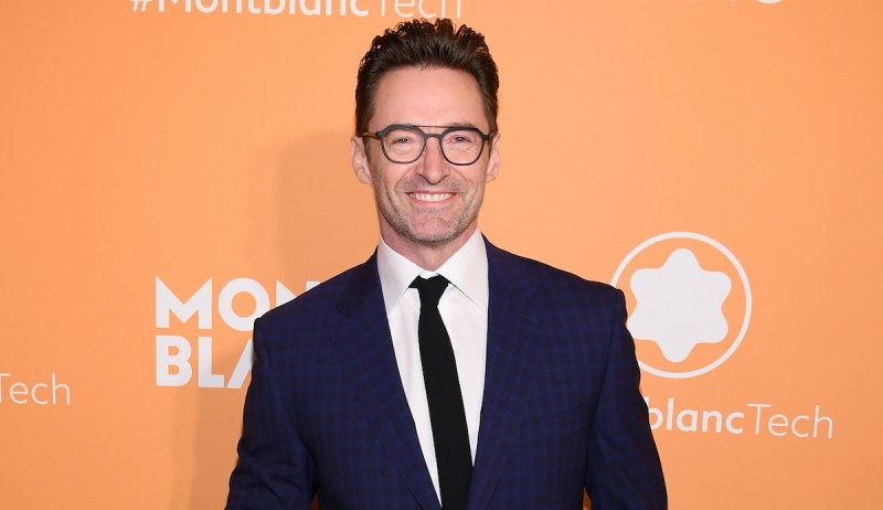 Hugh Jackman smiling in a navy suit against an orange background