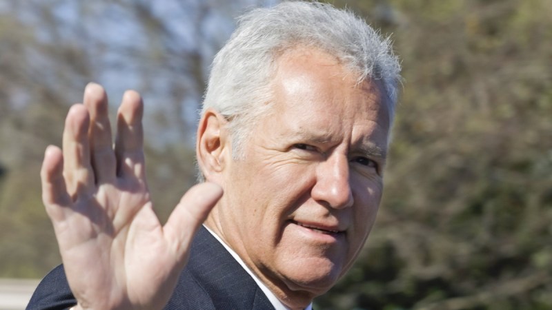 Alex Trebek walks outdoors wearing a dark suit with his hand raised to wave at photographers