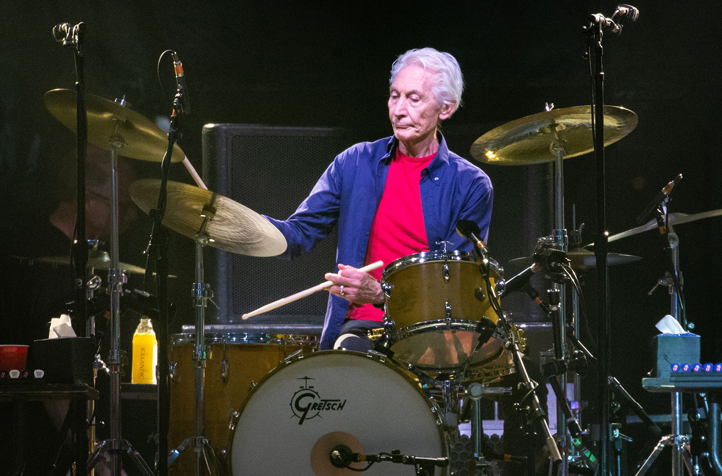Charlie Watts in a pink tee shirt and blue button down, playing drums on stage.