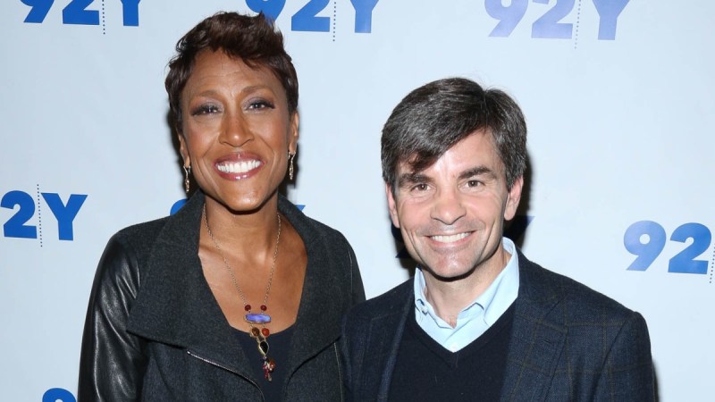 Robin Roberts, in all black, stands with George Stephanopoulos, in a dark blazer, pose together against a white background