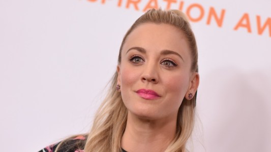 Kaley Cuoco wears her hair half up, half down against a white background while walking the red carpet