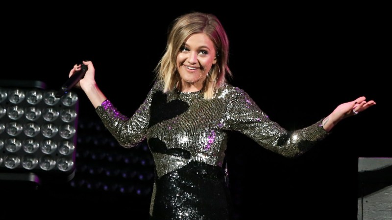 Kelsea Ballerini, wearing a silver dress festooned with black hearts, shrugs onstage