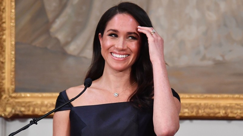 Meghan Markle smiles as she wears a black dress and pushes her hair back while giving a speech