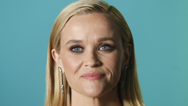 Reese Witherspoon stands before a teal background with her hair slicked behind her ears